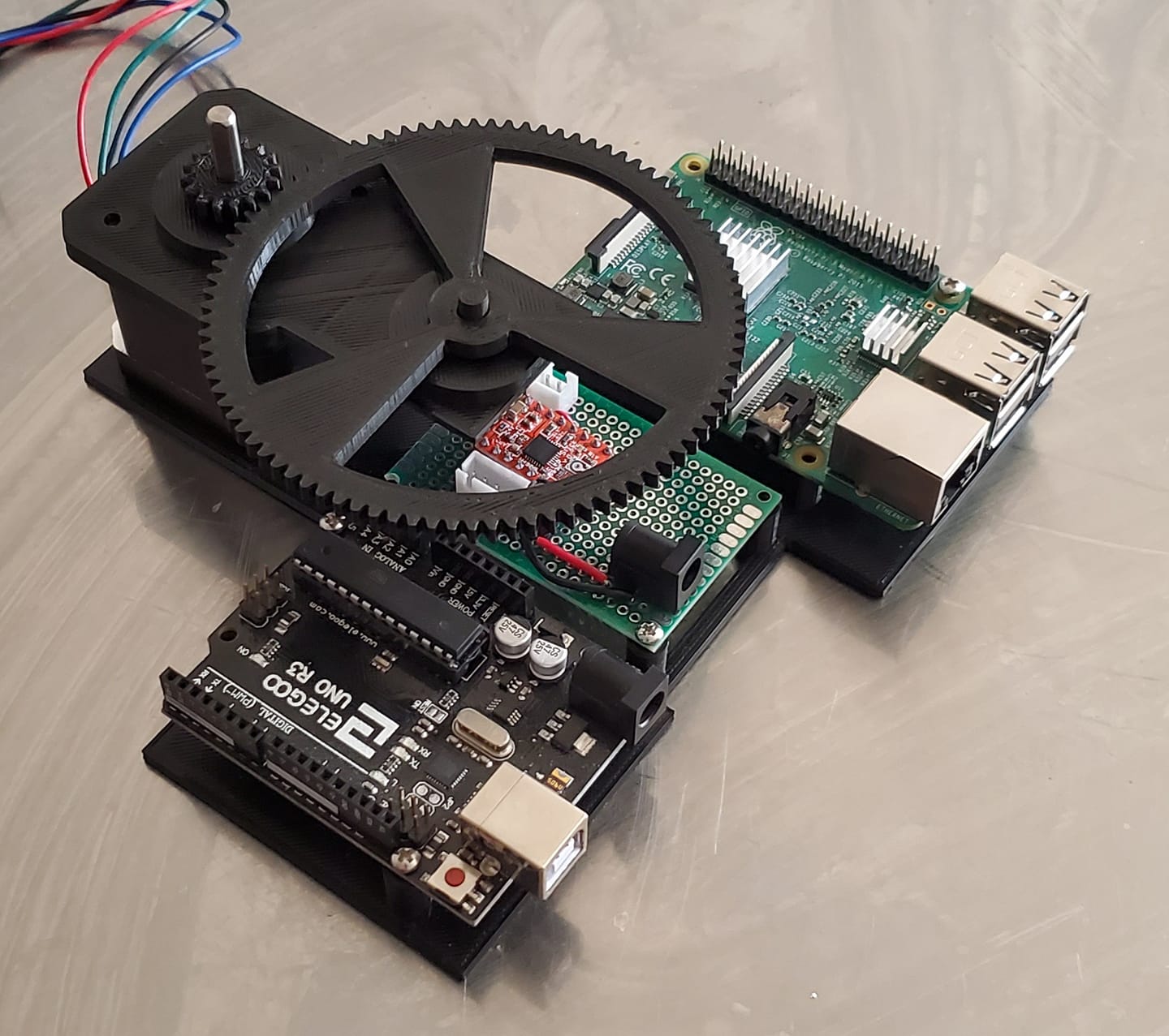 A device with a motor in center with gear on top, three spots on bottom of device that holds three circuits. Circuit on left is an arduino, middle is a soldering board with components soldered to it and right has a raspberry pie.